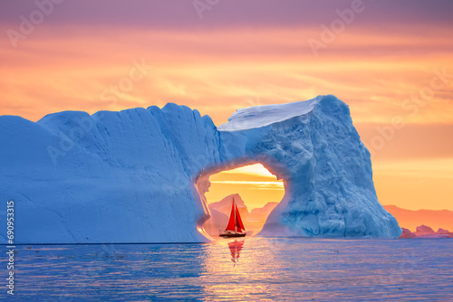 Beautiful landscape with little red sailboat cruising under a majestic iceberg arch during midnight sun season of polar summer in Greenland. Sail boat with red sails cruising among icebergs  Greenland