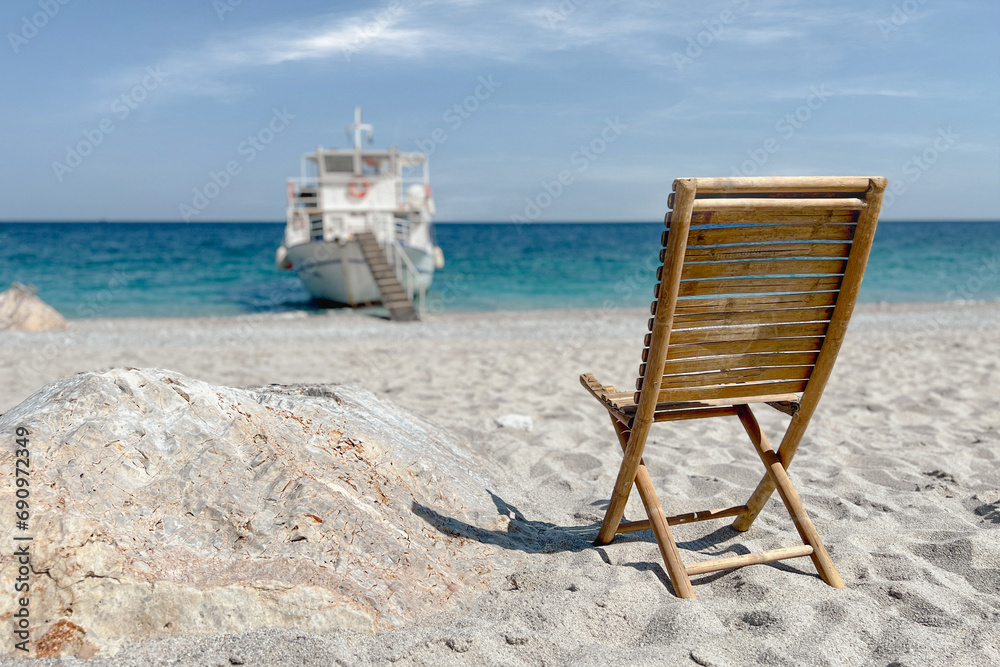 Empty wooden chair on sand beach faces towards boat and sea panorama. Summer travel concept.