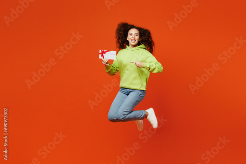 Full body young woman of African American ethnicity she wear green hoody casual clothes jump high hold point on gift certificate coupon voucher card for store isolated on plain red orange background.