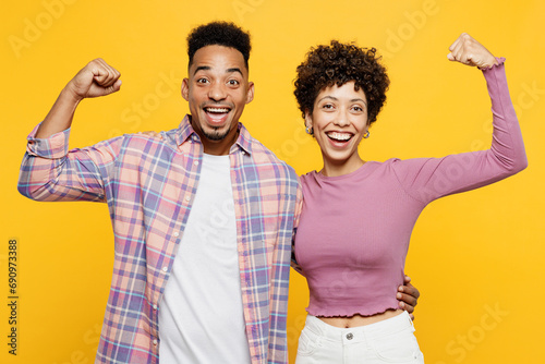 Young couple two friends family man woman of African American ethnicity wear purple casual clothes together show biceps muscles on hand demonstrating strength power isolated on plain yellow background