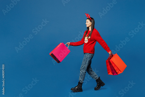 Merry young Latin woman wear red Christmas sweater fun deer horns on head posing hold paper package bags after shopping isolated on plain blue background. Happy New Year holiday, sale buy day concept. photo