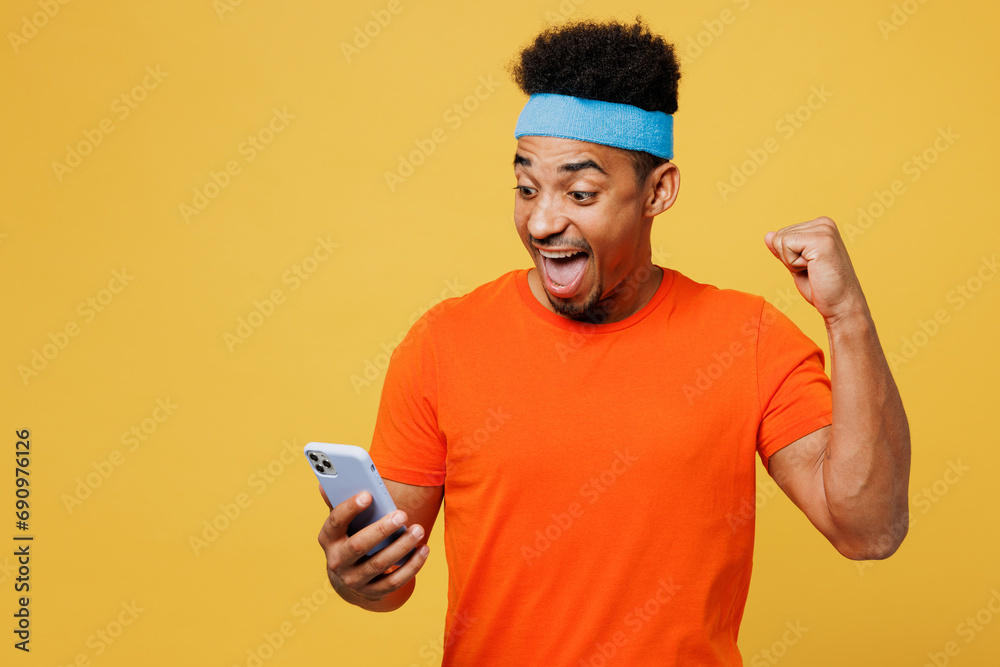 Young fitness trainer sporty man sportsman wear orange t-shirt hold use mobile cell phone do winner gesture spend time in home gym isolated on plain yellow background. Workout sport fit abs concept. Young fitness trainer sporty man sportsman wear orange t-shirt hold use mobile cell phone do winner gesture spend time in home gym isolated on plain yellow background. Workout sport fit abs concept.