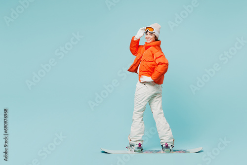 Full body sideways young woman wear warm padded windbreaker jacket hat ski goggles mask snowboarding look camera travel rest spend weekend winter season in mountains isolated on plain blue background photo