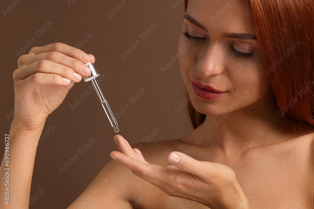 Fototapeta premium Beautiful young woman applying cosmetic serum onto her finger on brown background, closeup