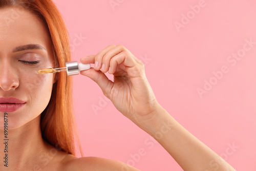 Beautiful young woman applying cosmetic serum onto her face on pink background, closeup. Space for text