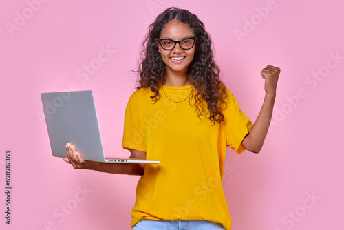 Young beautiful overjoyed Indian woman teenager holds laptop and enthusiastically waves hand after learning about admission to college or university on grant basis stands on pink background.