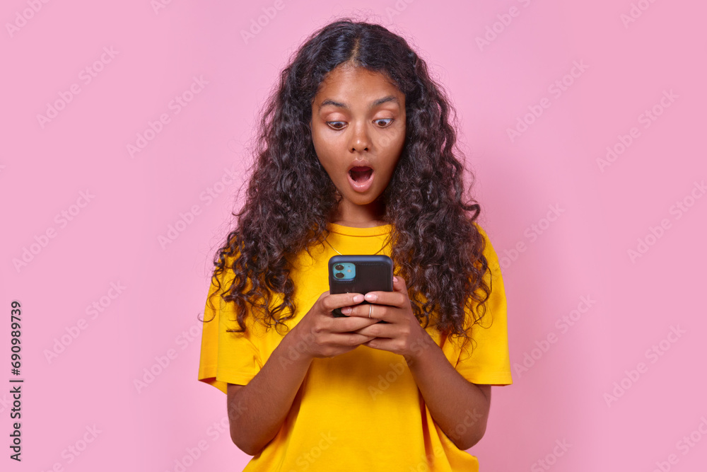 Young delighted beautiful Indian woman teen opens mouth in surprise reading message on mobile phone or seeing advertisement about upcoming concert of favorite band stands on pink background.