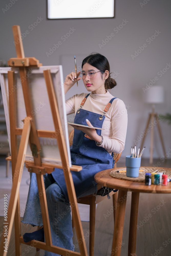 Young asian female artist touching glasses and looking on canvas to thinking idea painting artwork Young asian female artist touching glasses and looking on canvas to thinking idea painting artwork
