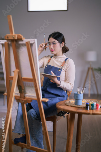 Young asian female artist touching glasses and looking on canvas to thinking idea painting artwork