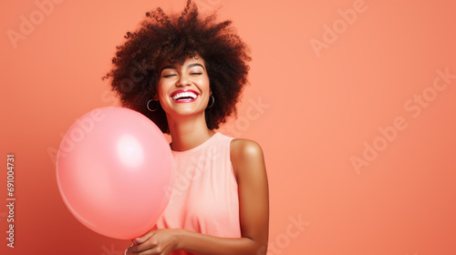 Joyful woman with an afro hairstyle, laughing and holding a pink balloon, wearing a light pink dress against a peach-colored background. © MP Studio