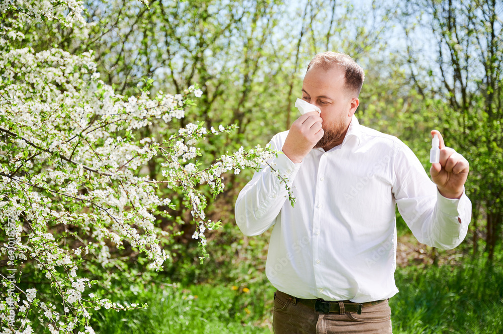 Man allergic using medical nasal drops, suffering from seasonal allergy in blossoming garden. Handsome man blowing nose with nasal handkerchief in front of blooming tree. Spring allergy concept. Man allergic using medical nasal drops, suffering from seasonal allergy in blossoming garden. Handsome man blowing nose with nasal handkerchief in front of blooming tree. Spring allergy concept.