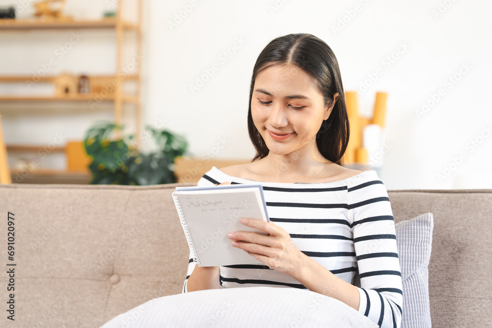 young attractive Asian woman smiling while reading a notebook, seated on a beige couch with a gray cushion. young attractive Asian woman smiling while reading a notebook, seated on a beige couch with a gray cushion.
