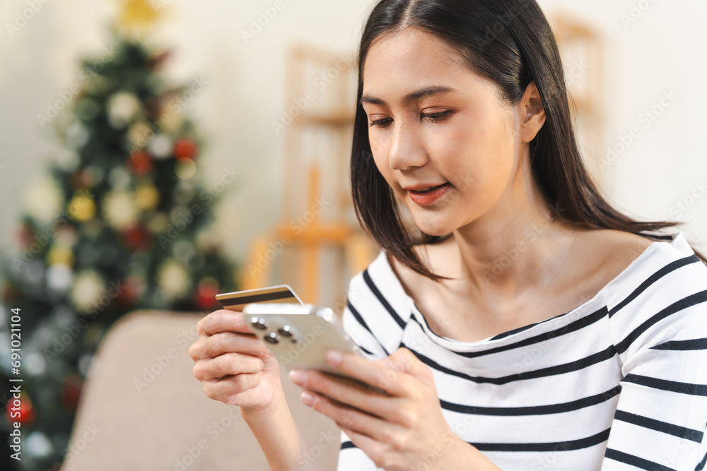 young, attractive Asian woman using a smartphone and holding a credit card, likely shopping online, with a Christmas tree in the background. young, attractive Asian woman using a smartphone and holding a credit card, likely shopping online, with a Christmas tree in the background.