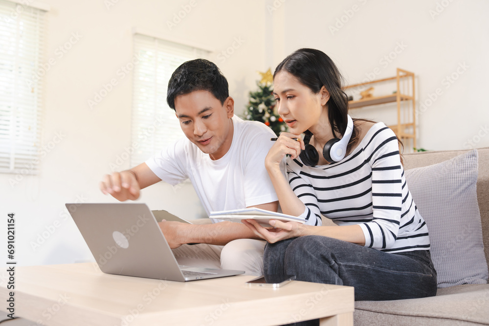 young attractive Asian woman and man couple sitting together, possibly making a Christmas gift list, with a festive tree in the background. young attractive Asian woman and man couple sitting together, possibly making a Christmas gift list, with a festive tree in the background.