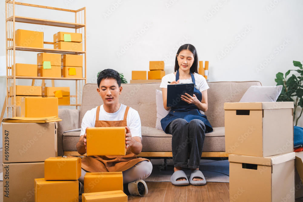 Asian man and woman in aprons work together, looking at paperwork and a laptop in a room filled with boxes. Asian man and woman in aprons work together, looking at paperwork and a laptop in a room filled with boxes.