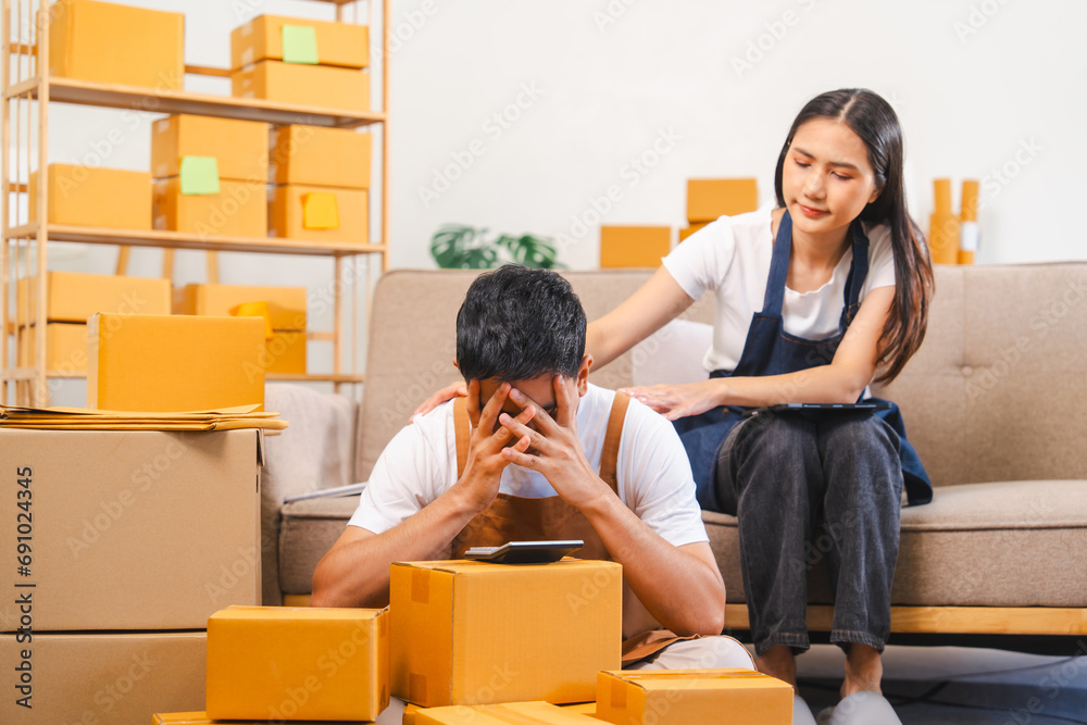 Asian man and woman look stressed, surrounded by boxes; the man holds a clipboard while the woman holds her head. Asian man and woman look stressed, surrounded by boxes; the man holds a clipboard while the woman holds her head.