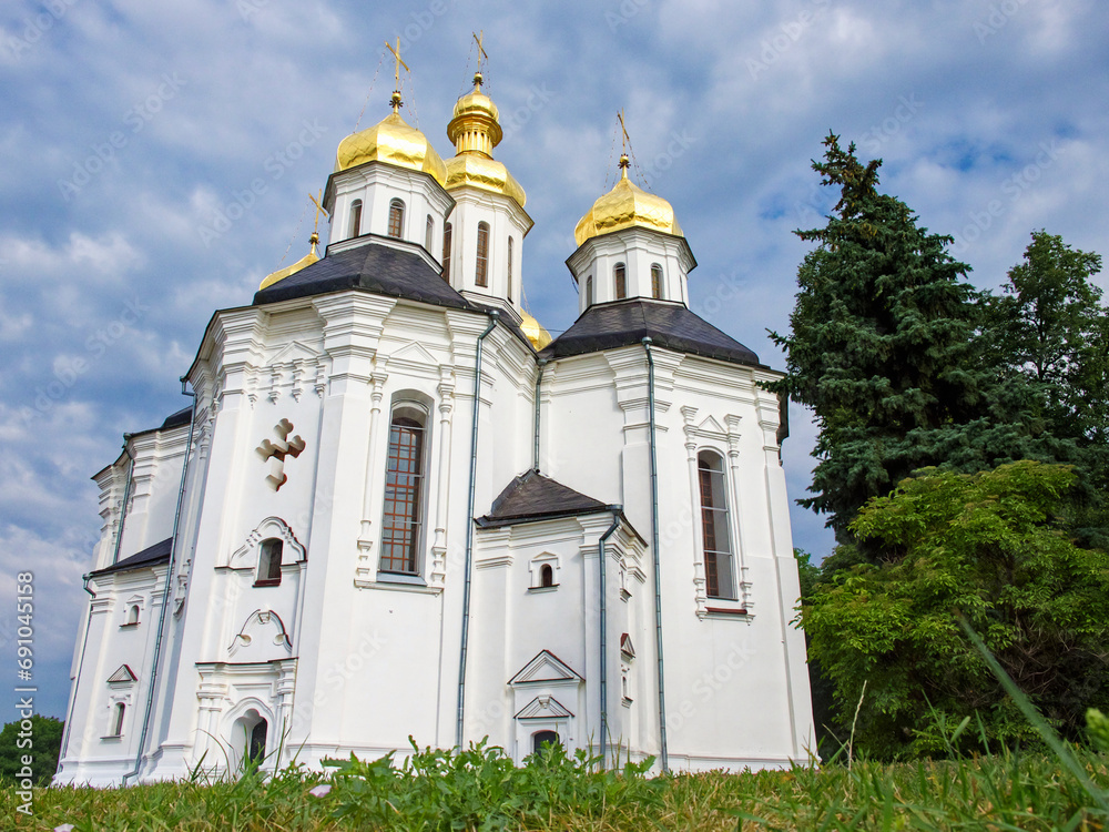The Orthodox Church of St. Catherine, a resplendent white church with gilded domes and crosses, as it stands in perfect harmony with the surrounding lush greenery and the tranquil blue sky.