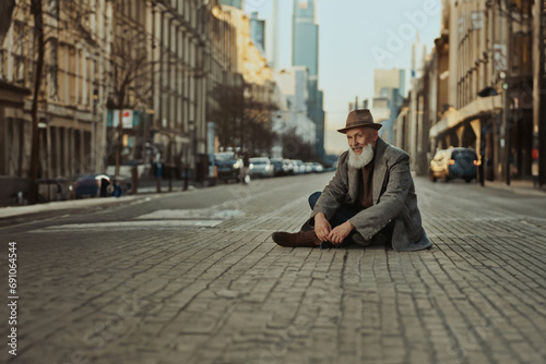  A desperate old man, Caucasian, gray beard sits on the ground on the street in the pedestrian zone in a city