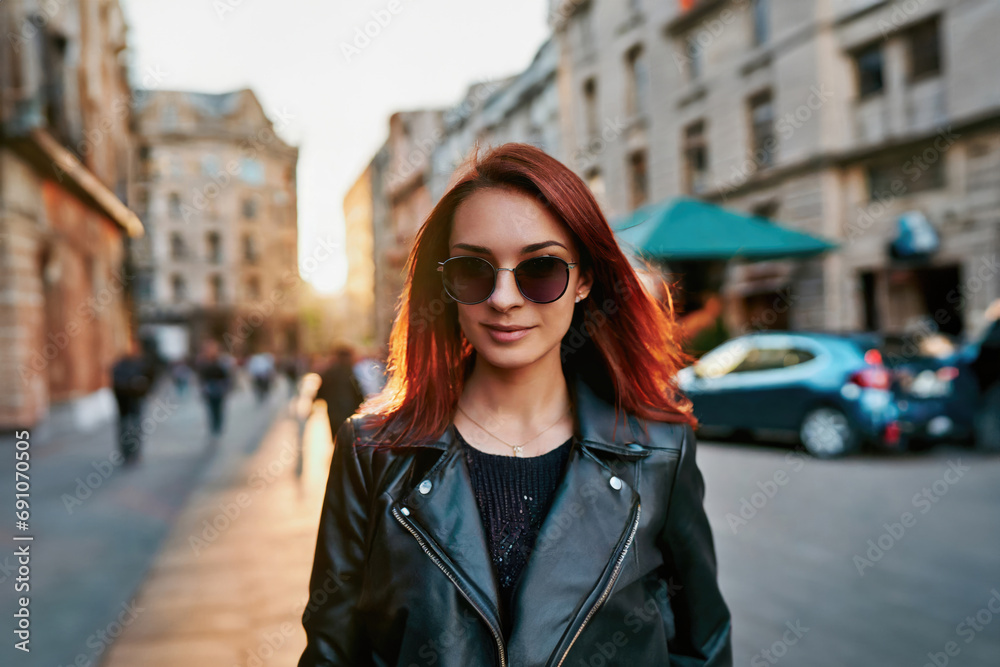  stylish young woman with red hair, wearing sunglasses and a black leather jacket, strolls on a lively urban street