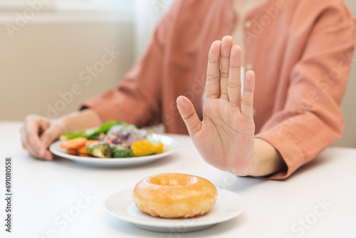 Diet concept, close up young woman, girl using hand push out, stop sweet donut, dessert or junk food on plate, choose green vegetables salad, eat low fat for good health. Female getting weight loss. photo