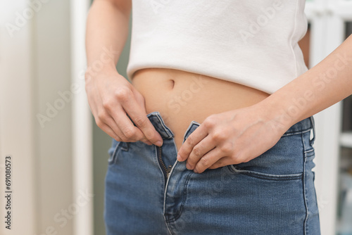 Excess weight gain, overweight, close up young woman, girl wearing fit trousers at home, hand trying to close zip up and button of jeans from fat around waist. Obese problem, health care concept. photo