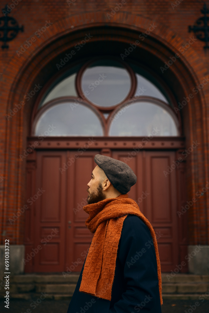 Naklejka premium A caucasian man with a beard standing in front of an old red wooden door wearing an orange scarf and a blue coat looking away from the camera.