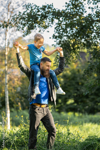 The little boy is very cheerful and laughs loudly as he is carried on his father's shoulders. A good dad entertains his son and plays with him during a walk in nature.