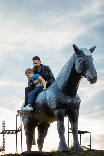 Portrait of a father and his little son on a big black horse against the background of the sky. Dad helps child climbs down the stairs photo