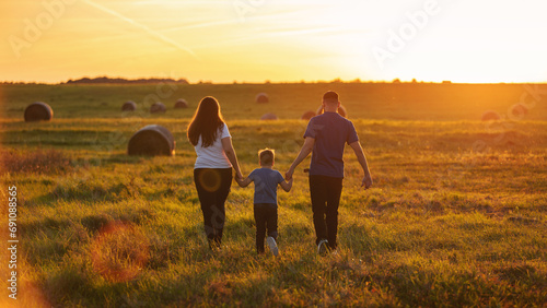 A young family from the back walking through the wide country fields holding hands in the rays of the golden sun  in the evening