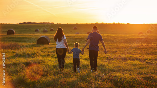 A young family from the back walking through the wide country fields holding hands in the rays of the golden sun, in the evening