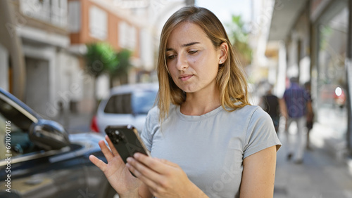Cool, young blonde woman in casual fashion, seriously engrossed in her smartphone outside on urban city street. concentrated young caucasian female texting, engrossed in her online lifestyle.