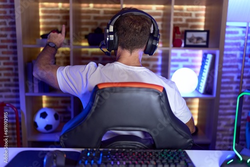 Middle age man with beard playing video games wearing headphones posing backwards pointing ahead with finger hand photo