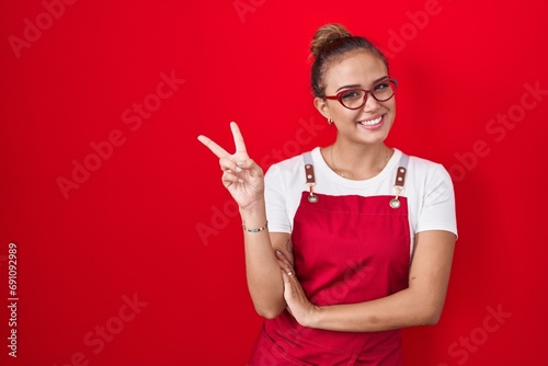 Young hispanic woman wearing waitress apron over red background smiling with happy face winking at the camera doing victory sign. number two.