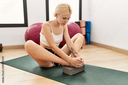 Young blonde woman sitting on yoga mat stretching legs at sport center