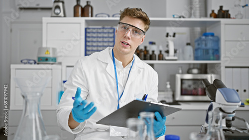 Attractive, smiling young caucasian male scientist, holding clipboard and passionately speaking about his groundbreaking research in the bustling lab