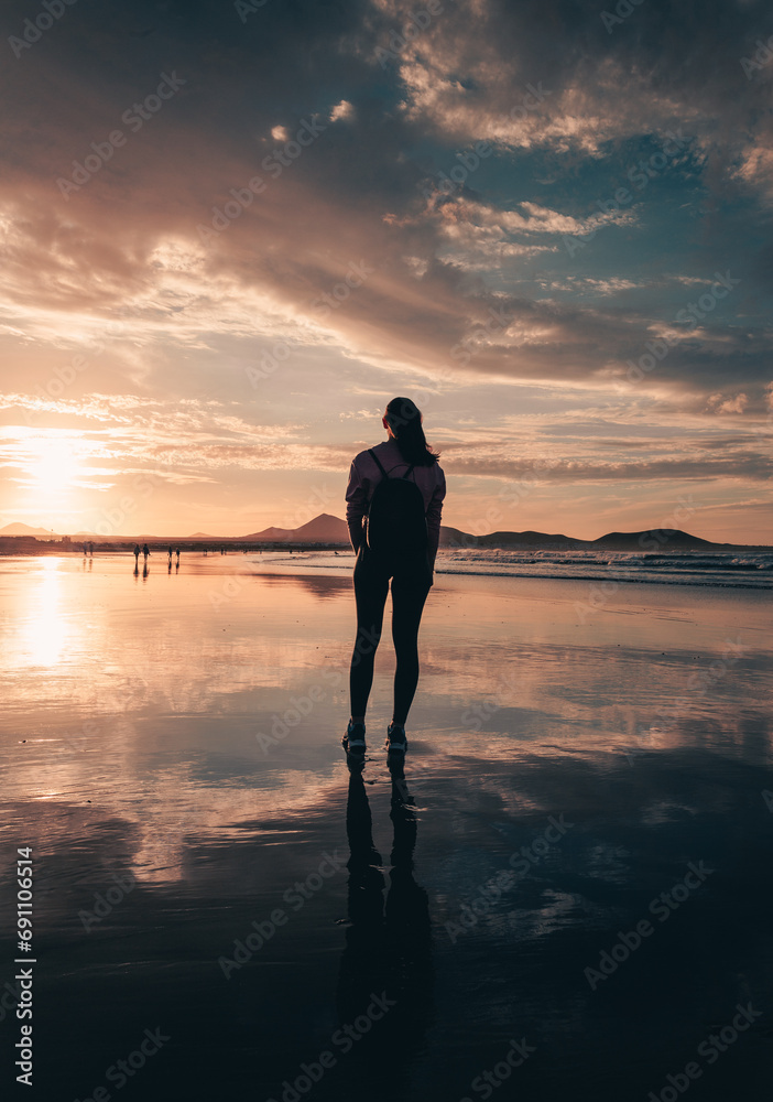 Vertical photo of free woman tourist on the beach during the sunset golden hour looking at the ocean. Woman on the beach with her reflection on wet sand - lifestyle concept.