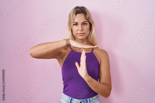Young blonde woman standing over pink background doing time out gesture with hands  frustrated and serious face