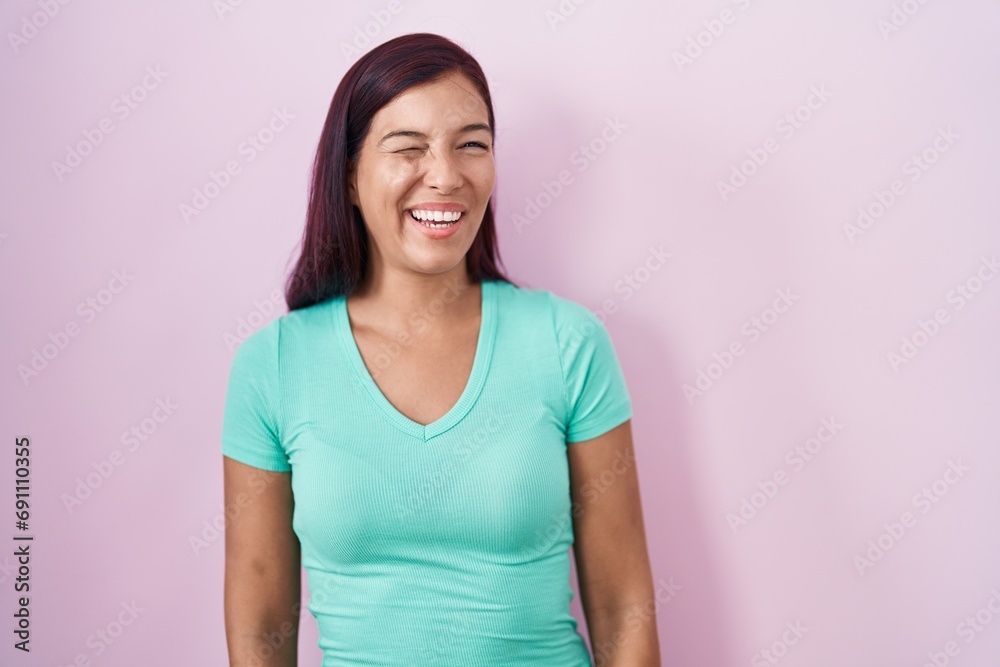 Fototapeta premium Young hispanic woman standing over pink background winking looking at the camera with sexy expression, cheerful and happy face.