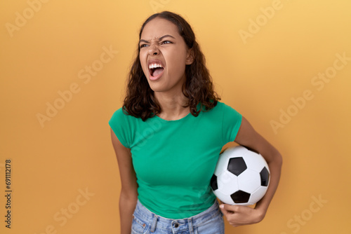 Young hispanic woman holding ball angry and mad screaming frustrated and furious, shouting with anger. rage and aggressive concept. © Krakenimages.com