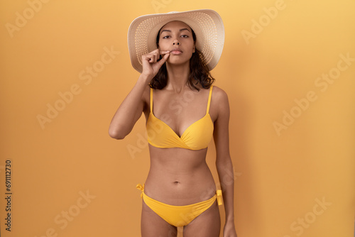 Young hispanic woman wearing bikini and summer hat mouth and lips shut as zip with fingers. secret and silent  taboo talking