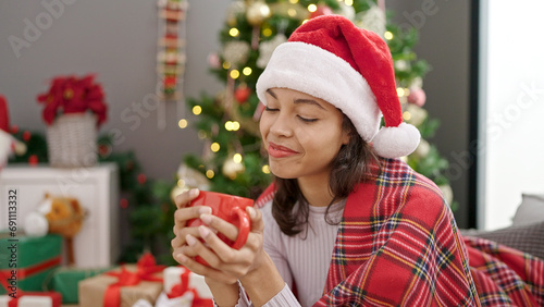 Young beautiful hispanic woman drinking coffee sitting by christmas decoration at home