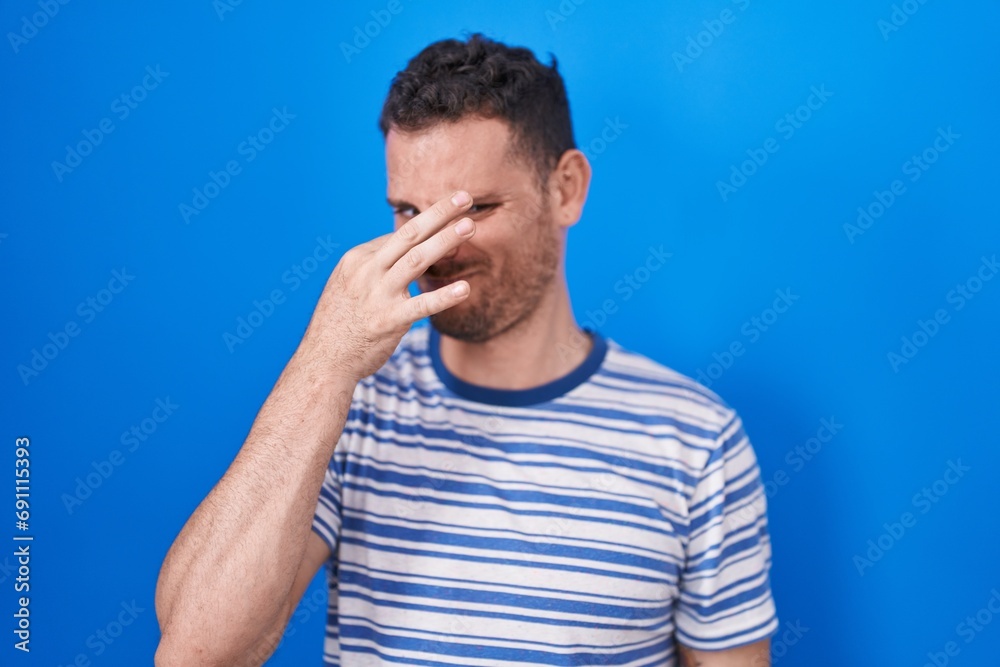 Young hispanic man standing over blue background smelling something stinky and disgusting, intolerable smell, holding breath with fingers on nose. bad smell Young hispanic man standing over blue background smelling something stinky and disgusting, intolerable smell, holding breath with fingers on nose. bad smell
