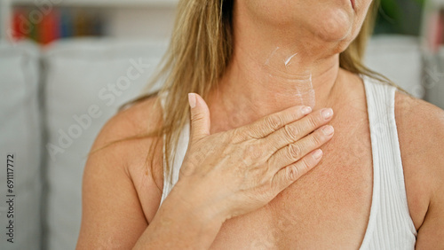 Middle age hispanic woman sitting on sofa applying lotion on neck at home photo