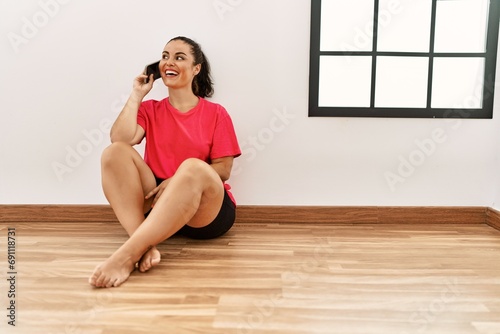 Young beautiful hispanic woman talking on smartphone sitting on floor at empty room
