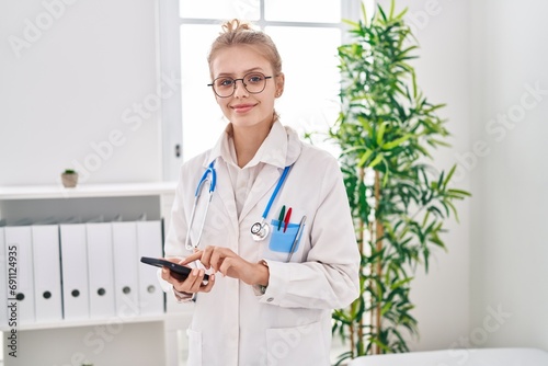 Young blonde woman doctor smiling confident using smartphone at clinic