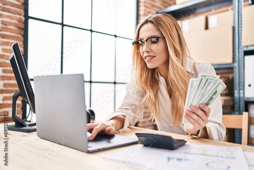 Young blonde woman ecommerce business worker using laptop holding dollars at office