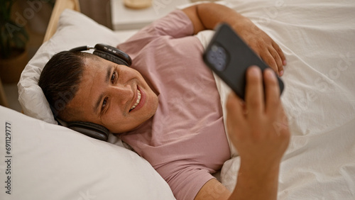 Cheerful young latin man joyously waving hello during a mobile video call indoors, lying content on his bedroom bed while using communication technology, house interior visible in the background