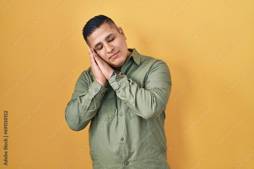 Hispanic young man standing over yellow background sleeping tired dreaming and posing with hands together while smiling with closed eyes.