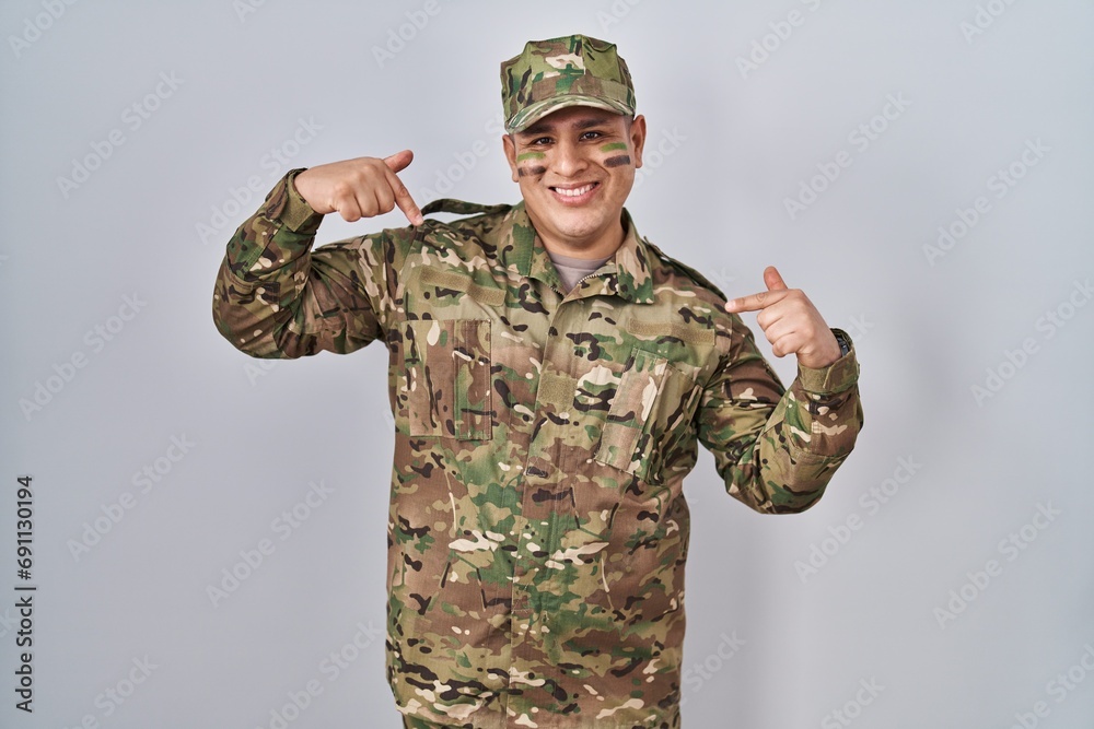 Hispanic young man wearing camouflage army uniform looking confident with smile on face, pointing oneself with fingers proud and happy. Hispanic young man wearing camouflage army uniform looking confident with smile on face, pointing oneself with fingers proud and happy.