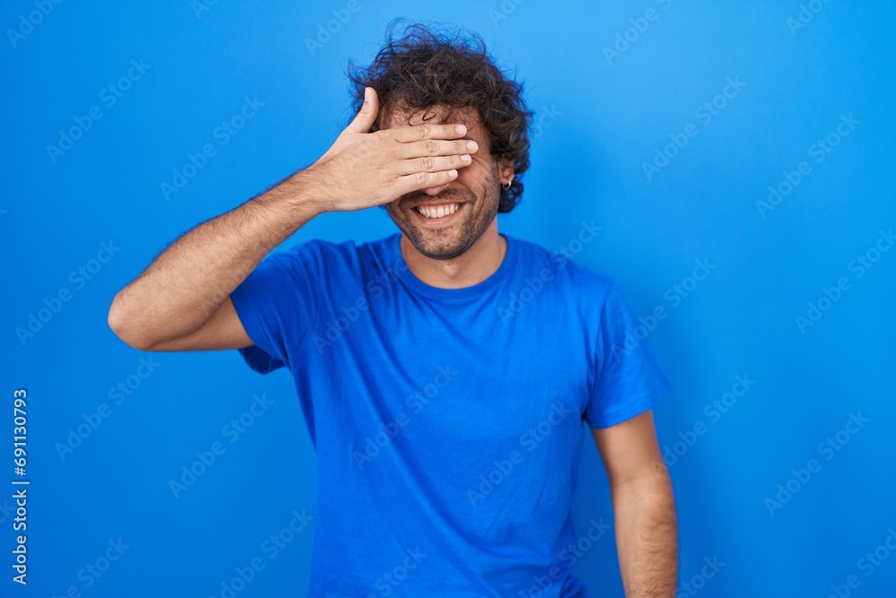 Hispanic young man standing over blue background smiling and laughing with hand on face covering eyes for surprise. blind concept. Hispanic young man standing over blue background smiling and laughing with hand on face covering eyes for surprise. blind concept.